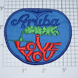May include: A blue, round embroidered patch with the text "Aruba I Love You" in red and white lettering. A green palm tree is depicted above the text.