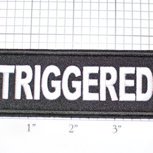 May include: A black fabric patch with white embroidered text that reads "TRIGGERED". The patch is approximately 3 inches wide and 1 inch tall.