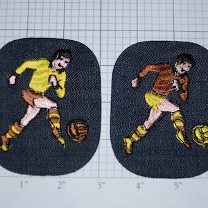 May include: Two embroidered patches featuring a soccer player kicking a ball. One player wears a yellow jersey, the other a brown jersey. Both patches have a dark blue background and are approximately 3 inches by 3 inches.