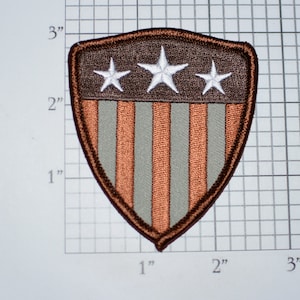May include: A brown and gray embroidered patch in the shape of a shield with a white star design and red and white stripes. The patch is approximately 2 inches wide and 3 inches tall.