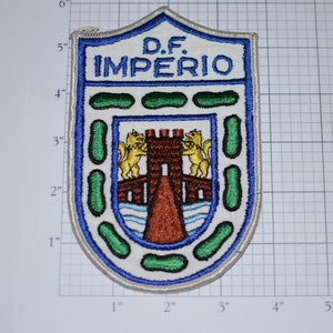 May include: A white embroidered patch with a blue border. The patch features a crest with a castle, two lions, and a bridge. The text "D.F. IMPERIO" is above the crest.