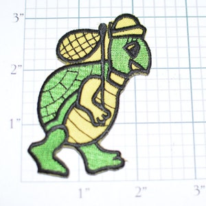 May include: Green and yellow embroidered patch of a turtle carrying a net on its back. The turtle is wearing a hat and walking to the right.
