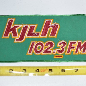 May include: A green fabric patch with the text "KJLH 102.3 FM" in red and yellow embroidered lettering.