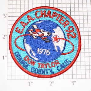 May include: A red, white, and blue embroidered patch with the text "E.A.A. CHAPTER 92" around a blue globe with a red airplane flying over it. The text "1976 DON TAYLOR ORANGE COUNTY, CALIF." is below the globe.