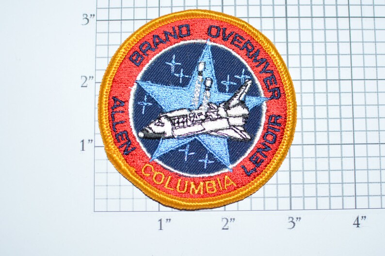 Op de afbeelding: Een geborduurde patch met een spaceshuttle, een ster en de tekst "BRAND OVERMYER ALLEN COLUMBIA LENDIR" in rood, wit en blauw.