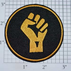 May include: A black and gold embroidered patch featuring a raised fist. The patch is circular and measures approximately 2 inches in diameter.