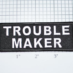 May include: Black and white embroidered patch with the text "Trouble Maker" in white lettering on a black background.