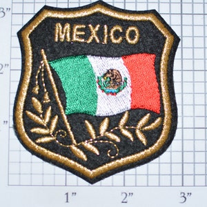 May include: A black embroidered patch with a gold border. The patch features the Mexican flag with the words "MEXICO" above it. The flag is waving and has a gold olive branch on either side.