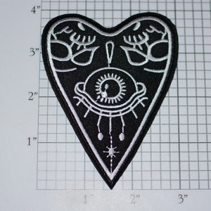 May include: Black felt patch in the shape of a heart with a white embroidered design of an eye with eyelashes and a star. The patch is approximately 4 inches tall.