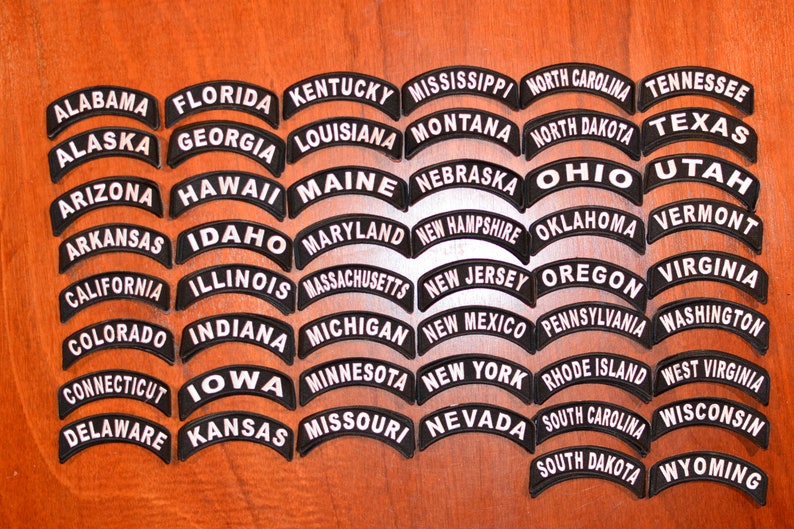 Puede incluir: Una colecci&oacute;n de parches bordados en blanco y negro, cada uno con el nombre de un estado de EE. UU. en un dise&ntilde;o curvo y arqueado. Los parches est&aacute;n dispuestos sobre una superficie de madera, mostrando los cincuenta estados.