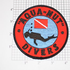 May include: A red and blue embroidered patch with the text "Aqua-Nut Divers" and a scuba diver silhouette. The patch features a red scuba diving flag with a white diagonal stripe.