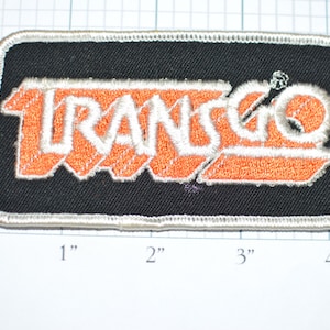 May include: An orange and white embroidered patch with the word "TRANSGO" in a stylized font. The patch is on a black background and has a white border.