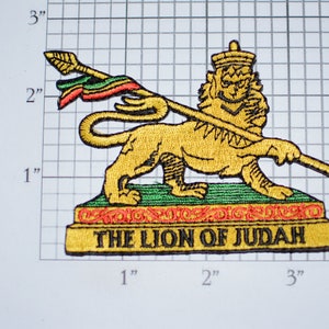 May include: Embroidered patch featuring a gold lion with a crown, holding a spear with a green, red, and yellow flag. The lion stands on a green and gold base with the text "THE LION OF JUDAH" in black.