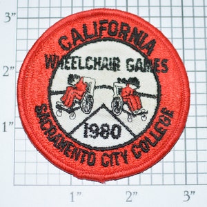 May include: A red, white, and black embroidered patch with the text "CALIFORNIA WHEELCHAIR GAMES SACRAMENTO CITY COLLEGE 1980". The patch features two black and white illustrations of people in wheelchairs.