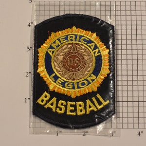 May include: A black felt patch with yellow embroidered text that reads "AMERICAN LEGION BASEBALL". The patch features a blue circle with a gold star and the letters "US" in the center. The circle is surrounded by a yellow wreath with orange accents.
