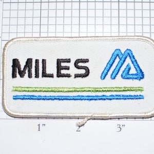 May include: White embroidered patch with the text "MILES" in black and a blue stylized mountain logo. The patch has two blue and one green embroidered lines.