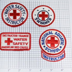 May include: Four embroidered patches with a red cross and blue text. The patches are for water safety instructors and swimmers. The patches are on a white grid background.