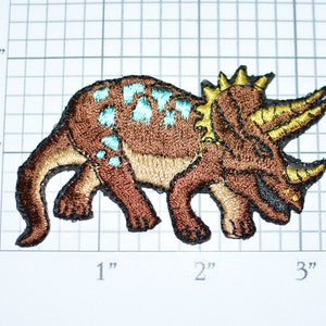 May include: Brown embroidered iron-on patch of a triceratops dinosaur with turquoise accents and gold horns. The dinosaur is walking with its tail curled behind it.