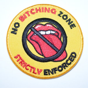 May include: A yellow embroidered patch with the text "No Bitching Zone Strictly Enforced" in red and black lettering. A red and black tongue with a diagonal line through it is in the center of the patch.