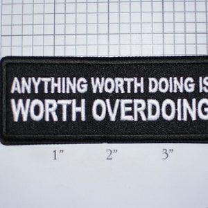 May include: Black embroidered patch with white text that reads "Anything worth doing is worth overdoing".