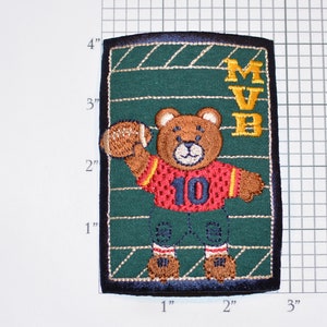 May include: Embroidered patch featuring a brown teddy bear wearing a red and white football jersey with the number 10. The bear is holding a brown football and is standing on a green field with the letters MVB in yellow.