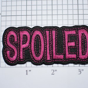 May include: Embroidered patch with the word "SPOILED" in bright pink, outlined in black. The patch is approximately 2 inches long and 1 inch tall. The patch has a black border and is on a grid background.