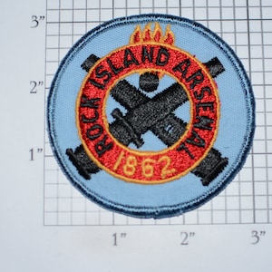 May include: A blue and red embroidered patch with the text "Rock Island Arsenal 1862" and a crossed cannon design. The patch has a yellow border and a flame design above the text.