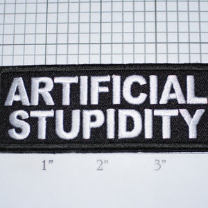 May include: A rectangular black patch with white embroidered text that reads "ARTIFICIAL STUPIDITY". The patch is approximately 4 inches long and 1 inch tall.