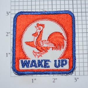 May include: A red and blue embroidered patch with a white rooster in the center and the text "WAKE UP" below. The rooster is facing right and has a red comb and wattle.
