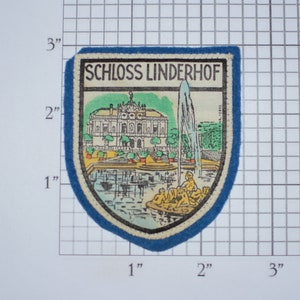 May include: A blue and white embroidered patch with the text "SCHLOSS LINDERHOF" and a colorful illustration of a palace with a fountain in front of it.