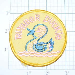 May include: A yellow embroidered patch with a blue rubber duckie swimming in pink waves. The text "Rubber Duckie" is embroidered in pink around the duckie.
