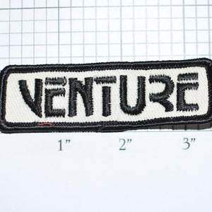 May include: Rectangular embroidered patch with the word "VENTURE" in black, block letters. The patch has a white background and a black border. The patch measures approximately 3 inches long and 1 inch high.