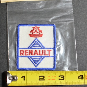 May include: A blue and white embroidered patch with the word "RENAULT" in red lettering. The patch features a stylized logo of a ship's wheel with a diamond shape in the center.