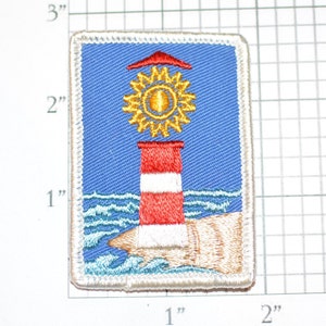 May include: A white, red, and yellow embroidered patch of a lighthouse with a sun on top, set against a blue background with white waves at the bottom. The patch is 2 inches by 2 inches.