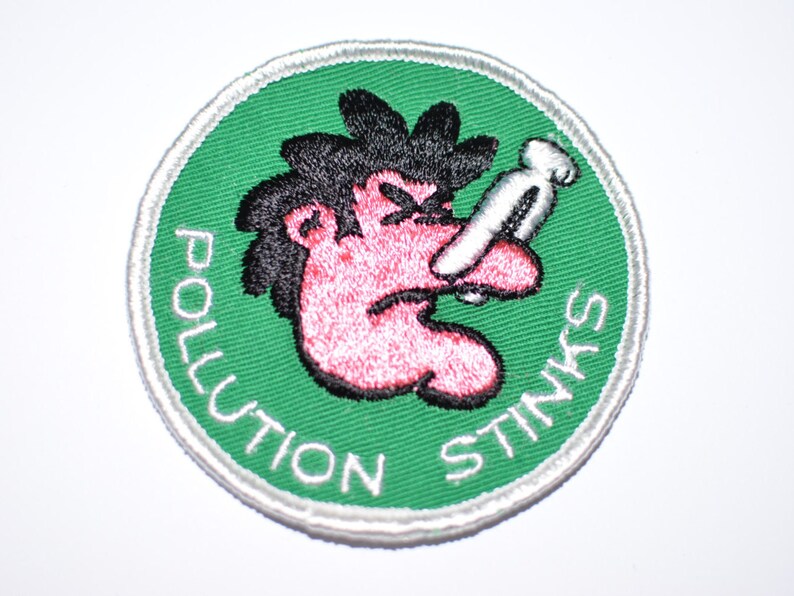 May include: A green and white circular patch with a cartoon illustration of a person with a red face and black hair holding their nose. The text "Pollution Stinks" is written around the edge of the patch.