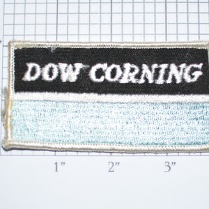 May include: A white and black embroidered patch with the text "DOW CORNING" in white lettering on a black background. The patch has a white border and a light blue background.