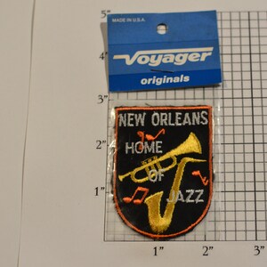 May include: A black embroidered patch with orange stitching. The patch features a trumpet and saxophone with musical notes and the text "New Orleans Home of Jazz".