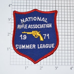 May include: A blue and red embroidered patch with the text "NATIONAL RIFLE ASSOCIATION 1971 SUMMER LEAGUE" and a gold silhouette of a handgun.