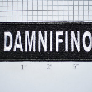 May include: A black embroidered patch with white lettering that reads "DAMNIFINO". The patch is approximately 3 inches wide and 1 inch tall.