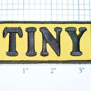 May include: A yellow fabric patch with black embroidered letters spelling "TINY".