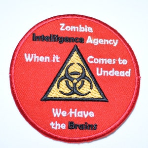 May include: Red round embroidered patch with a white border. The patch features a black triangle with a yellow biohazard symbol inside. The text around the triangle reads "Zombie Intelligence Agency When it Comes to Undead We Have the Brains".