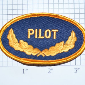 May include: Oval-shaped patch with the word "PILOT" in gold lettering on a navy blue background. The patch is bordered with gold stitching and features gold laurel leaves. The patch measures approximately 3 inches wide and 2 inches tall.