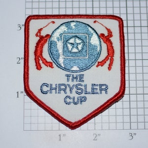 May include: White embroidered patch with red border. The patch features a blue globe with a white star in the center. Two red figures are swinging golf clubs above the globe. The text "THE CHRYSLER CUP" is embroidered in blue below the globe.