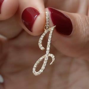 May include: A gold-colored pendant in the shape of the letter "J", encrusted with small, sparkling gemstones. The pendant is held up against a blurred background. The gemstones are clear and reflect light.