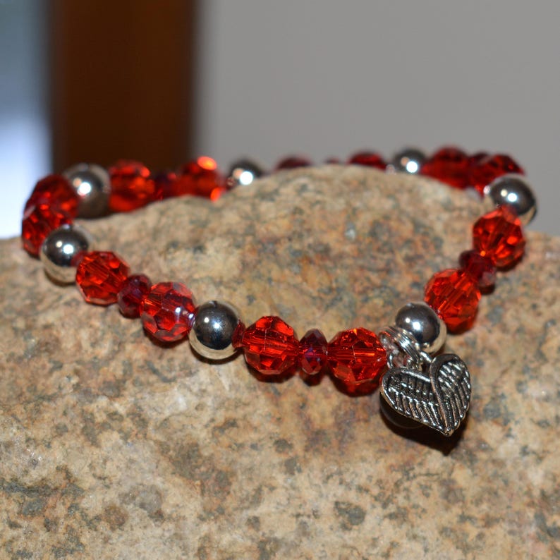 Puede incluir: Una pulsera roja con cuentas, detalles plateados y un dije de coraz&oacute;n. La pulsera presenta cuentas rojas facetadas y cuentas plateadas lisas. El dije de coraz&oacute;n tiene un dise&ntilde;o de ala detallado. La pulsera se muestra sobre una superficie de piedra.