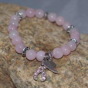 Op de afbeelding: Een roze rozenkwarts armband met zilveren kralen en een bedel met een roze lint met strass. De armband ligt op een grijze rotsachtige achtergrond.
