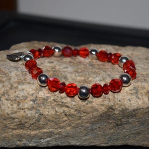Puede incluir: Una pulsera con cuentas rojas facetadas y cuentas de metal plateado. La pulsera est&aacute; adornada con un peque&ntilde;o dije de hoja plateada. La pulsera se exhibe sobre una superficie de piedra texturizada de color marr&oacute;n claro.