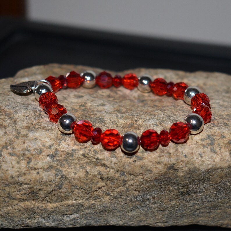 Puede incluir: Una pulsera con cuentas rojas facetadas y cuentas de metal plateado. La pulsera est&aacute; adornada con un peque&ntilde;o dije de hoja plateada. La pulsera se exhibe sobre una superficie de piedra texturizada de color marr&oacute;n claro.