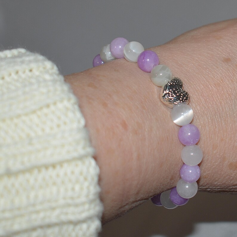 May include: A purple and white beaded bracelet with a silver heart charm. The bracelet is made with round beads and is worn on a person's wrist.