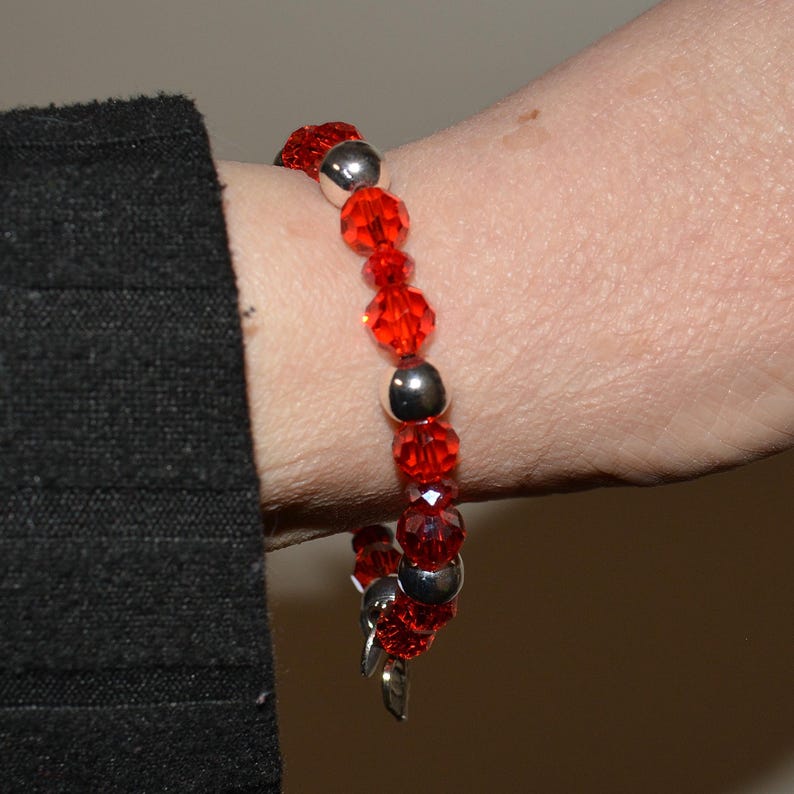 Puede incluir: Una pulsera de cuentas rojas con detalles plateados. La pulsera presenta cuentas rojas facetadas y cuentas plateadas lisas y redondas. La pulsera se lleva en la mu&ntilde;eca.